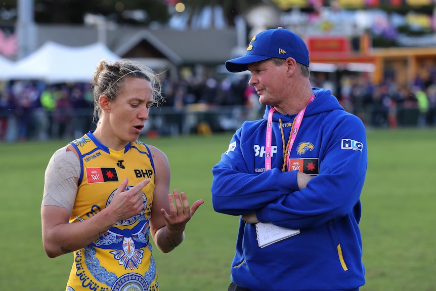 A man in a blue cap and jumper chats to a female football player wearing yellow