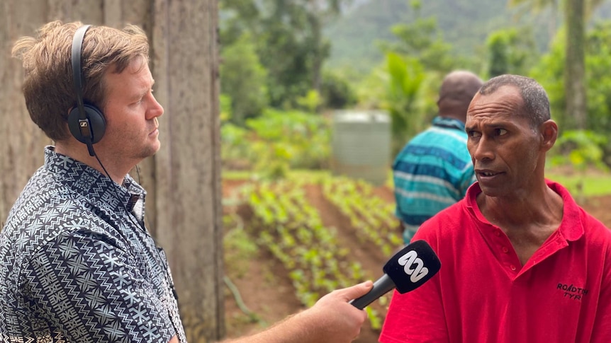 Fred interviews a Fijian man in a field using headphones and microphone.