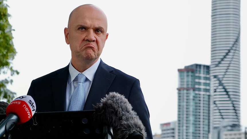 Doctor John Gerrard frowns at a lecturn packed with microphones with the Brisbane skyline in the background. 