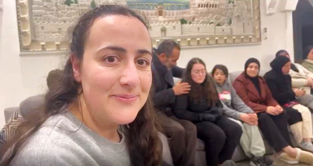 A woman smiles at the camera, as several people sit on a sofa behind her