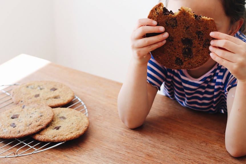A child holds a giant choc chip cookie in front of their face, with a wire rack of cookies nearby.