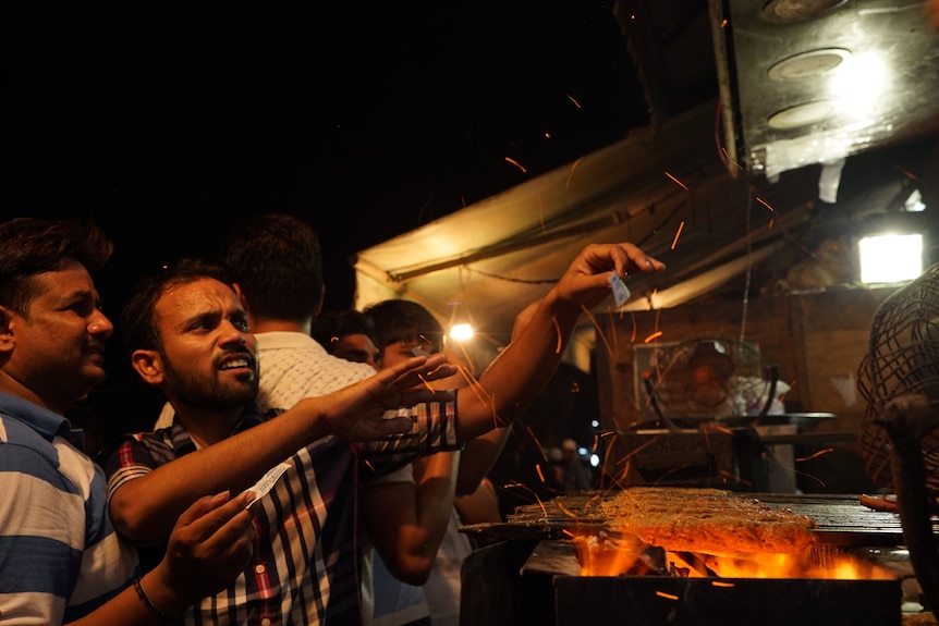 Two men standing in front of a street stall barbecue try to get the attention of the server.