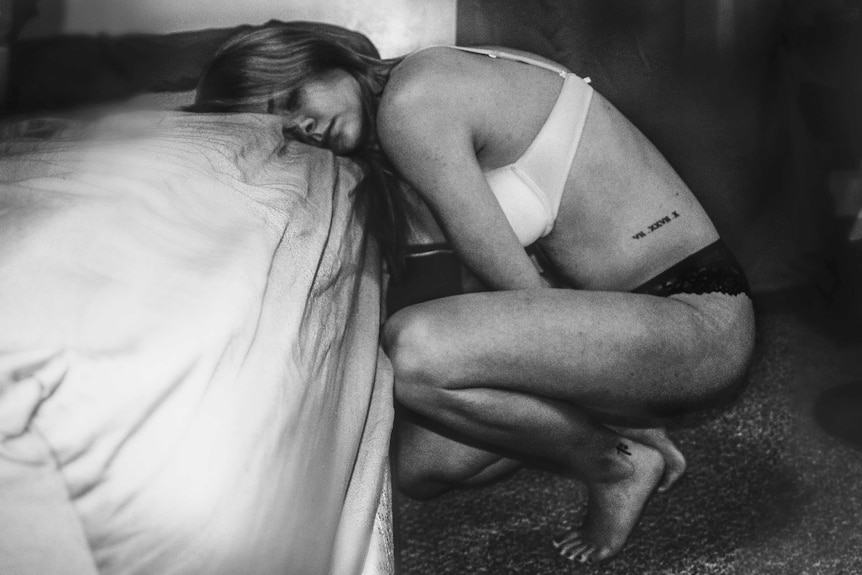 Woman in bra and panties curled up in pain alongside a bed 