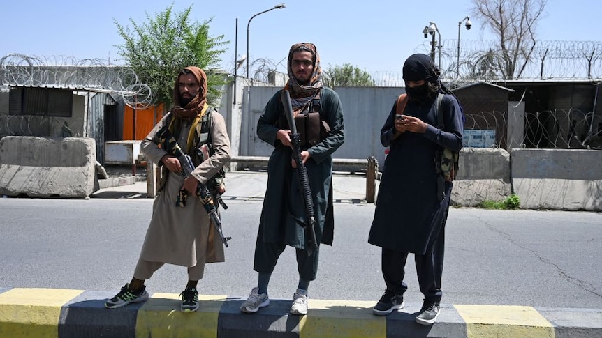 three young taliban fighters stand on guard on the road, two carrying guns and one on his phone
