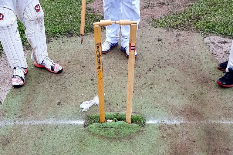 A cricket wicket with leg stump, off stump and bails in place, while a person in cricket whites holds the other stump.