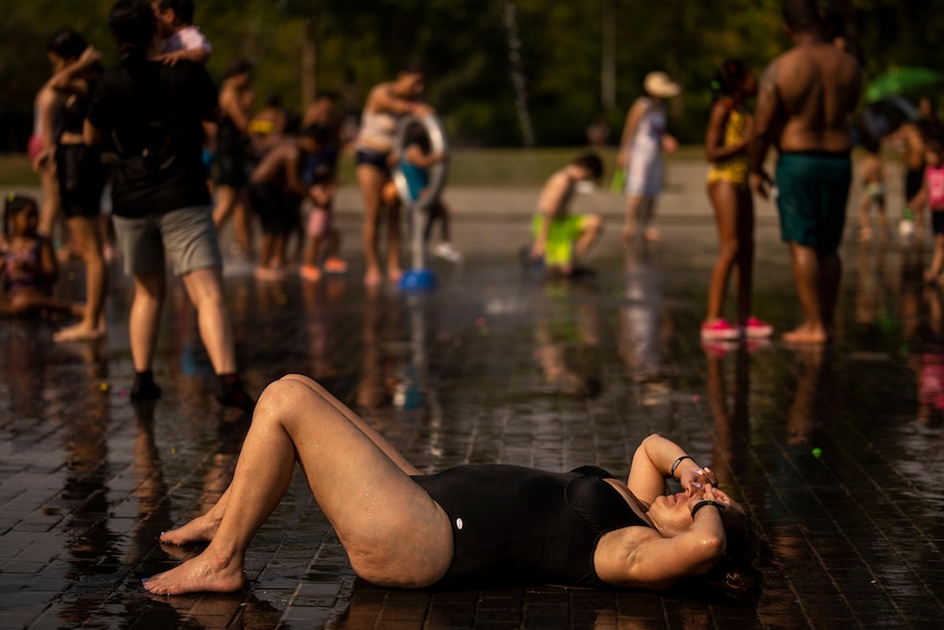 A woman lies on the ground of an urban water park, shielding her face from the sun as she cools down. 