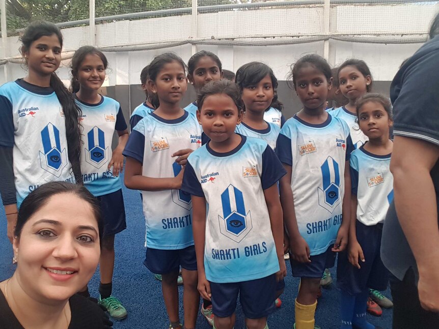 About 10 girls in soccer uniform look into Rica's camera as she takes a selfie on the training pitch.