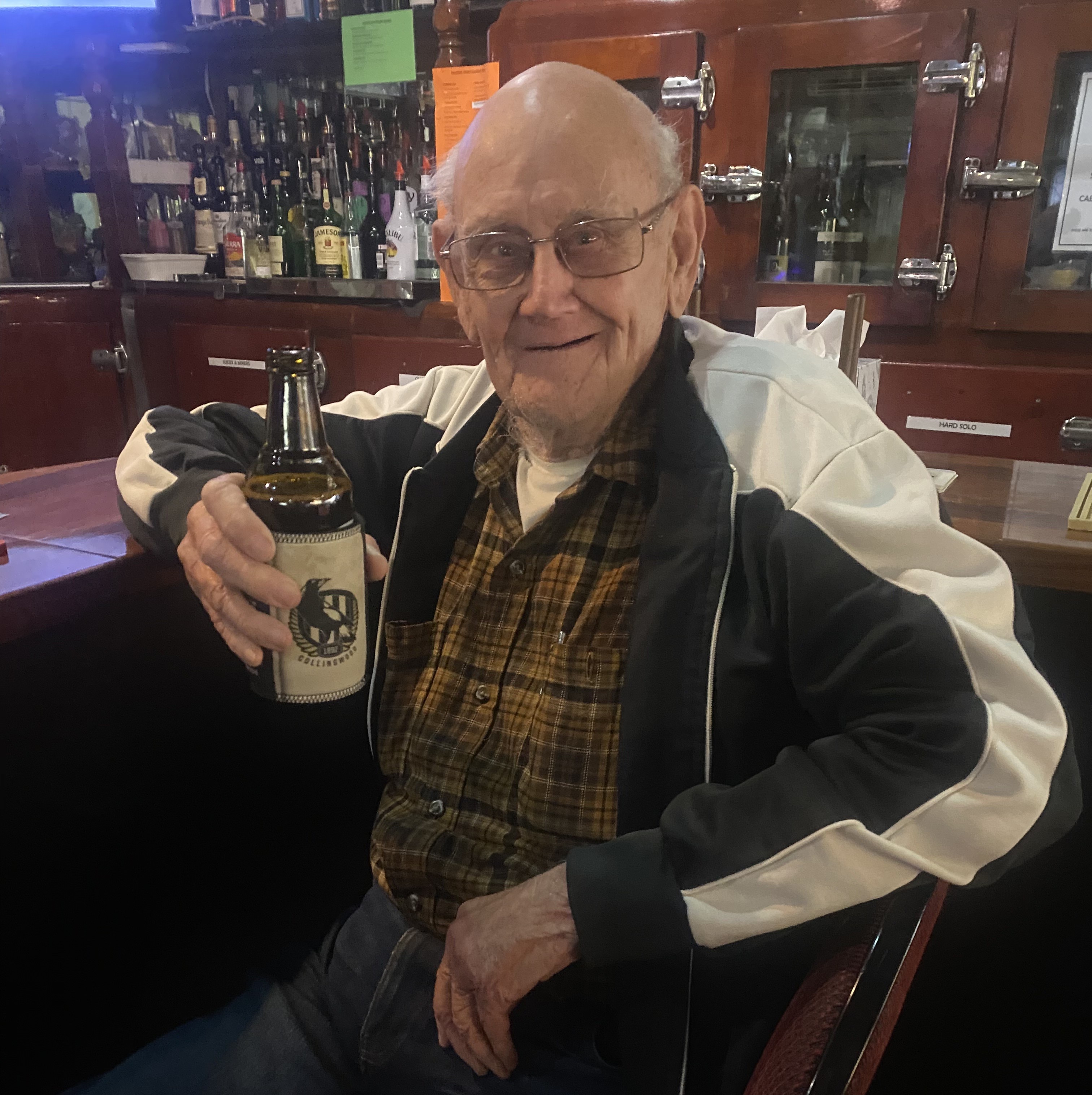 Harry, a man sitting on a stool next to a bar, holding a bottle of beer in a stubby holder and smiling at the camera 