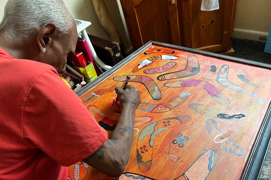 A man bends over a large Indigenous artwork in a frame and applies the finishing touches