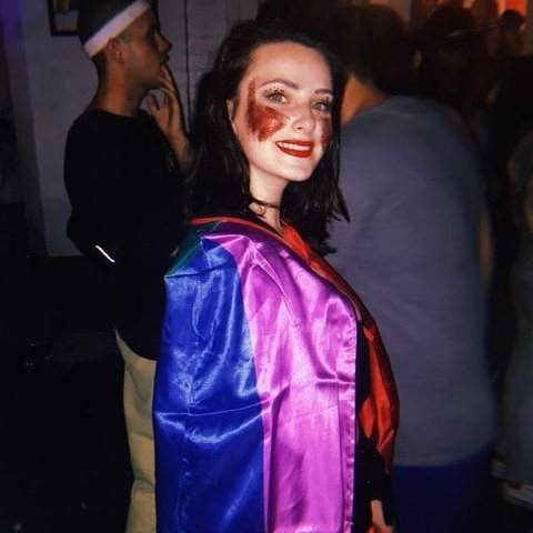 A photo of a young woman, Jordyn, wearing a rainbow flag around her neck and red glitter on her cheeks.