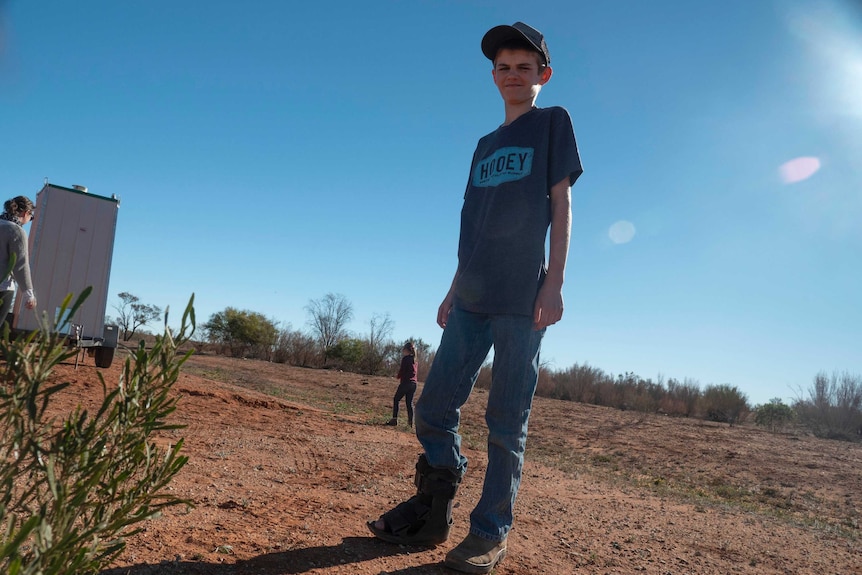 A young boy stands on red dirt, with a leg in a cast.
