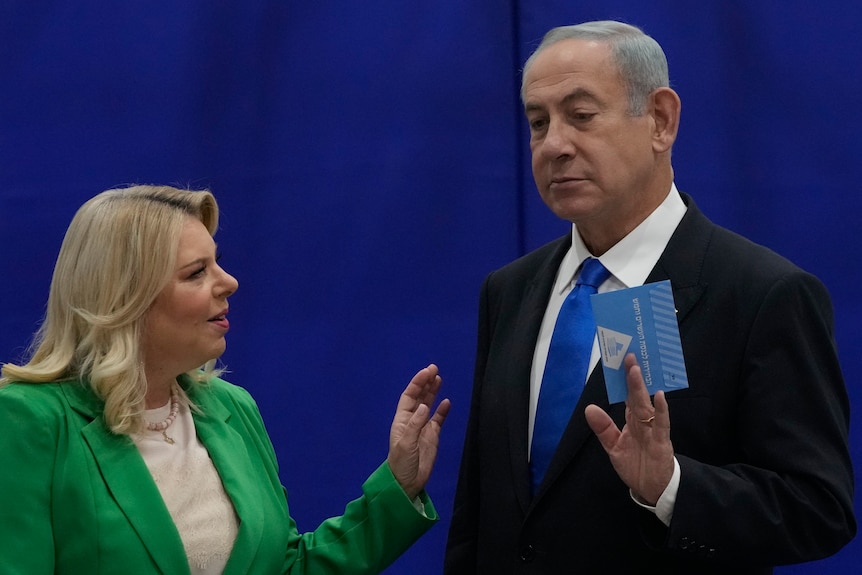 A suited man holds a voting ballot in his left hand while a woman in a light green coast moves to put her hand on his shoulder. 