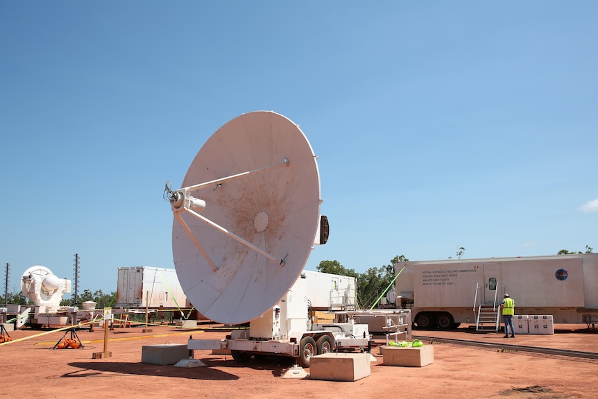 A satellite and some boxes outside on red dirt underneath a blue sky.
