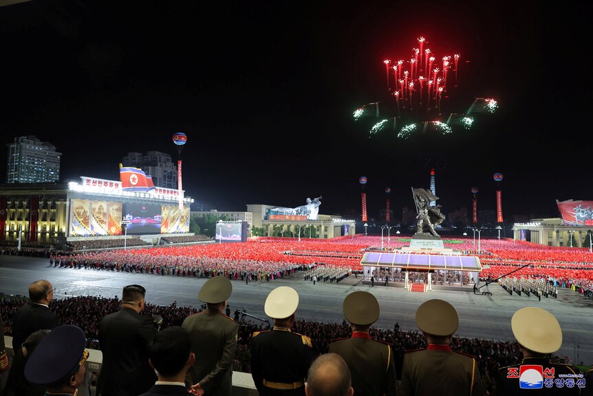 A fireworks display over soldiers is watched by men in military hats in raised seating