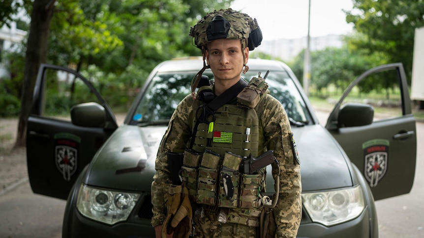 A man dressed in an army uniform with a helmet in front of a parked car. 