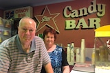 An elderly man and woman stand next to a popcorn machine in a movie theatre candy bar.
