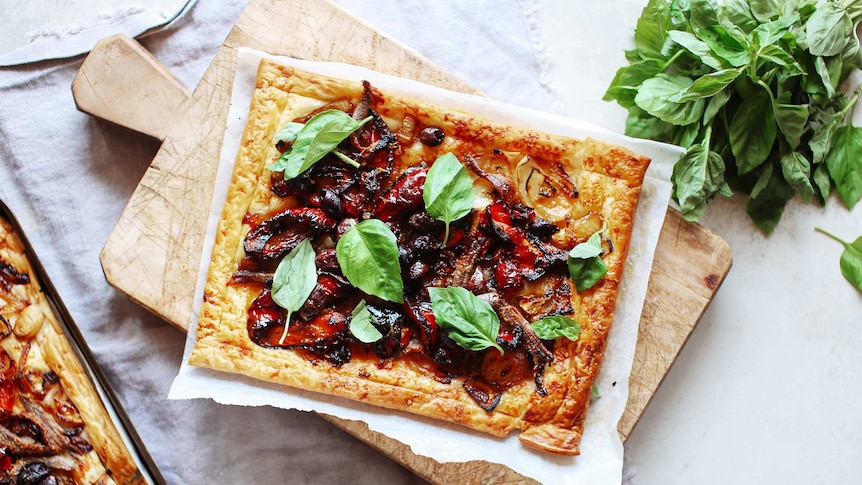 A puff pastry tart topped with capsicum, onions, anchovies and fresh basil sits on a chopping board, a recipe ready for lunch.