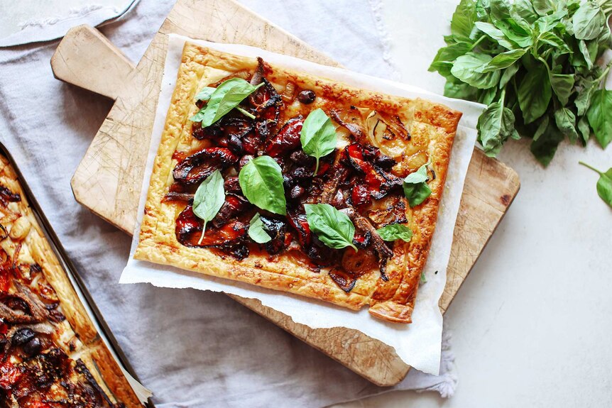 A puff pastry tart topped with capsicum, onions, anchovies and fresh basil sits on a chopping board, a recipe ready for lunch.