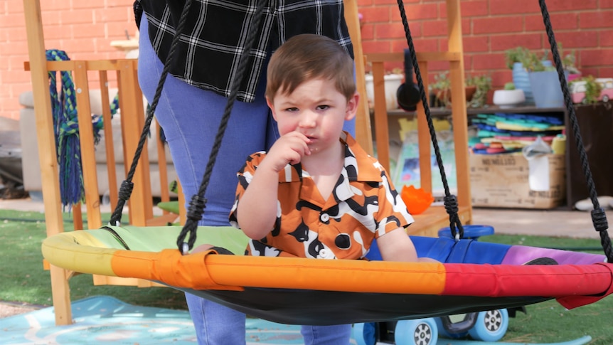 A one year old boy with brown hair and a orange patterned shirt sits on a swing being pushed by his mum in his backyard 