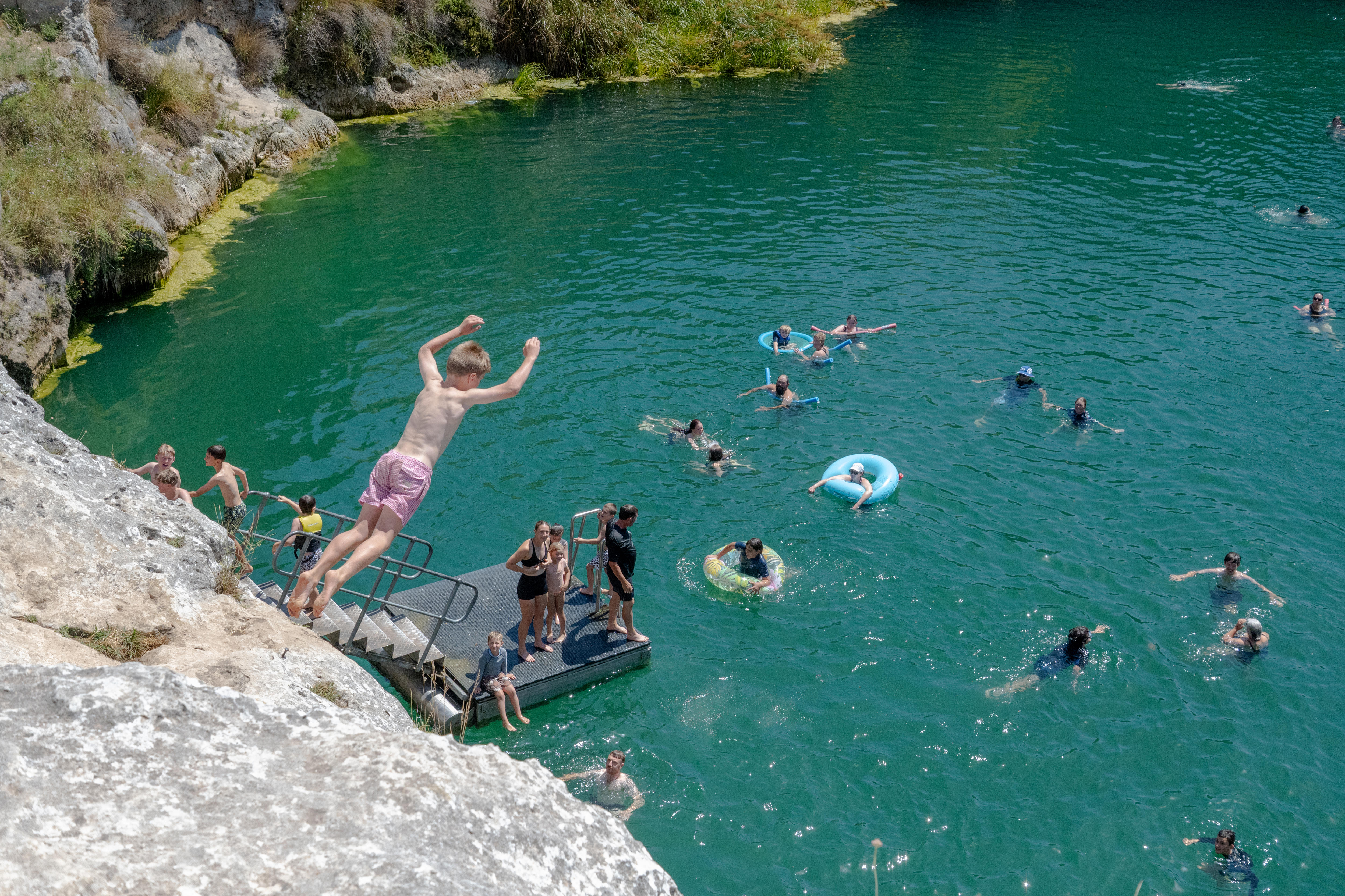 Un niño con shorts de baño rosas salta desde un acantilado rocoso a un lago lleno de agua con nadadores debajo