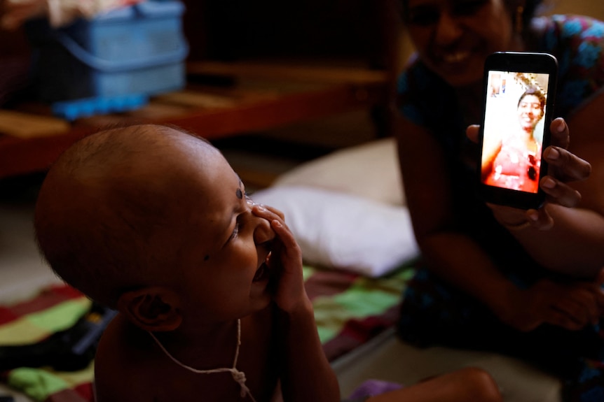 a young boy laughs and holds his hand up to his face as he looks at video phone call image of his mother on a phone