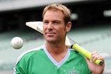 Shane Warne, in a green cricket shirt, stands with a bat and ball in the MCG.