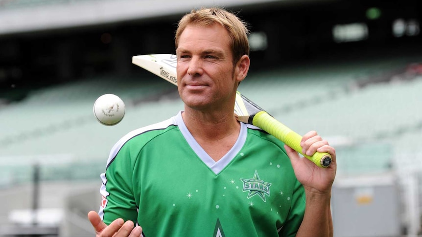 Shane Warne, in a green cricket shirt, stands with a bat and ball in the MCG.