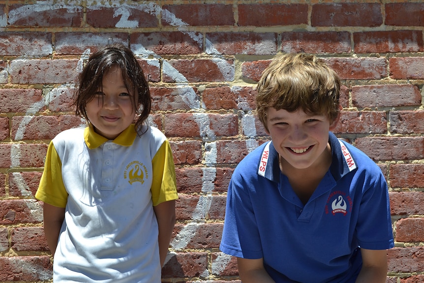 Colour photo of primary school students Genevieve and Darcy posing on a sunny day in front of brick wall.