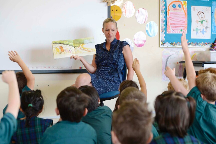 A female teacher holding a picture book open in front of students  who are sitting on the floor in front of her.