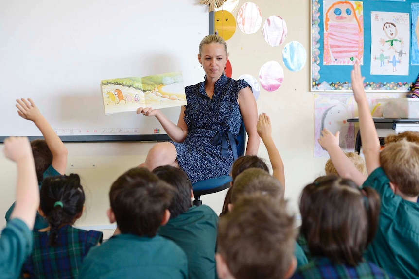 A female teacher holding a picture book open in front of students  who are sitting on the floor in front of her.