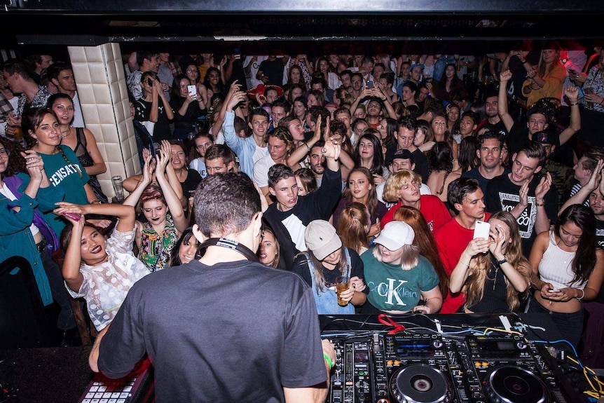 A crowd applauds a DJ playing music at a nightclub.