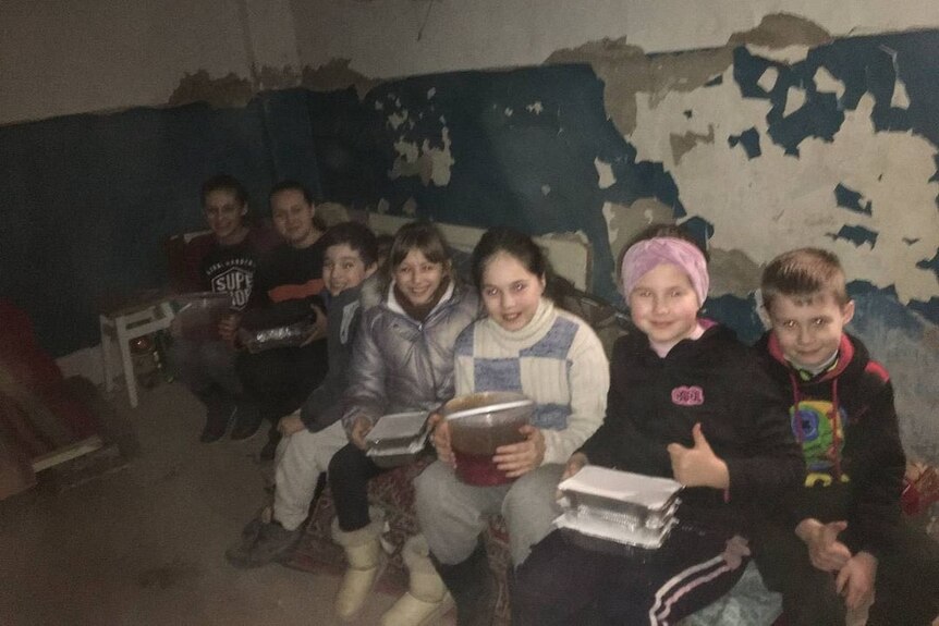 Children in a bomb shelter hold packaged up meals in their laps.