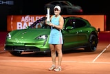 Ash Barty stands smiling with a trophy in front of a green Porsche.