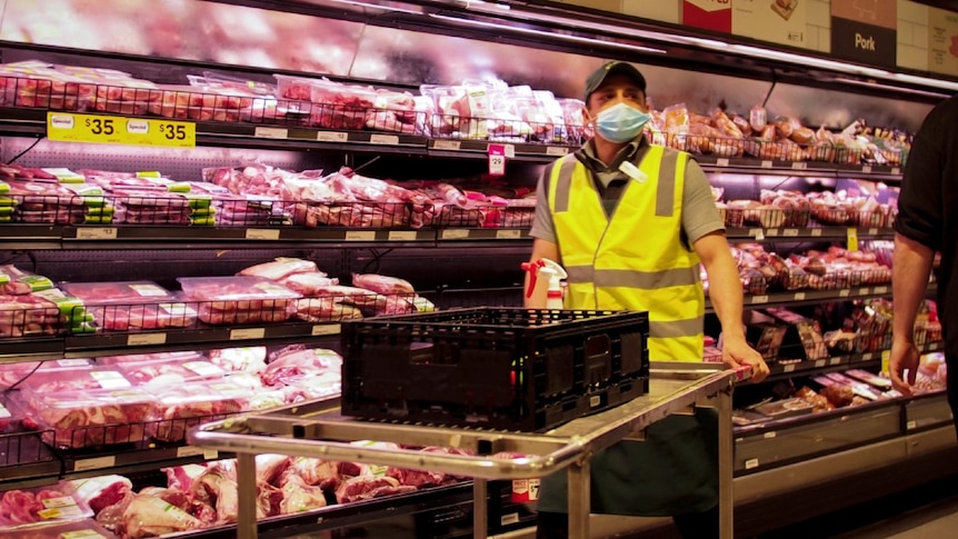 A worker in a high-vis vest wearing a blue surgical mask next to rows of red meat in an aisle in Woolworths.
