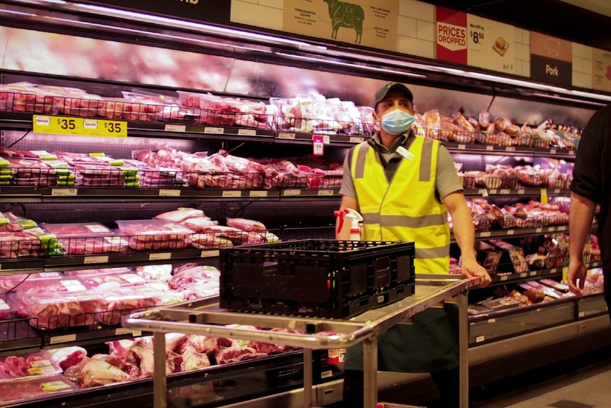 A worker in a high-vis vest wearing a blue surgical mask next to rows of red meat in an aisle in Woolworths.