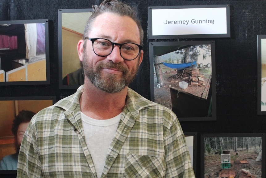 A man in a checkered shirt stands in front of photos on a black background. 