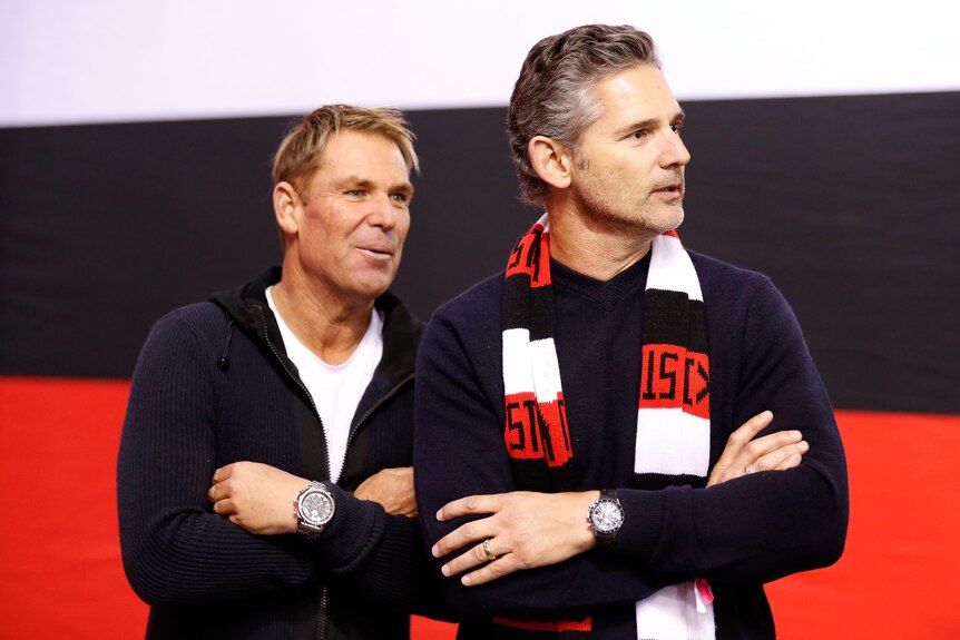 Two men stand together, one of which wears a black, white and red scarf
