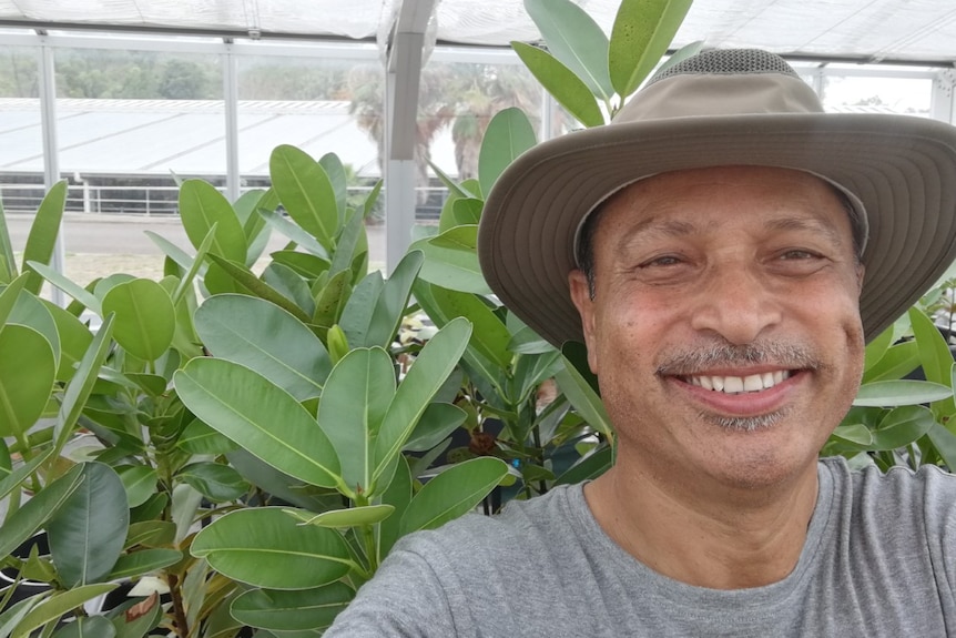 A man taking a selfie and smiling, there are trees with large leaves in the background.