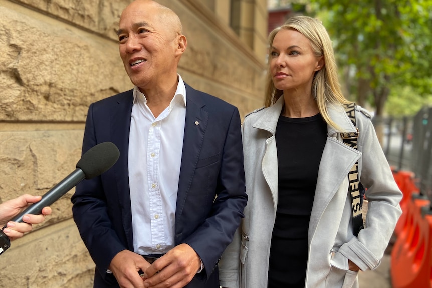 A blond woman and a bald man in a suit talking to the media next to the courthouse building