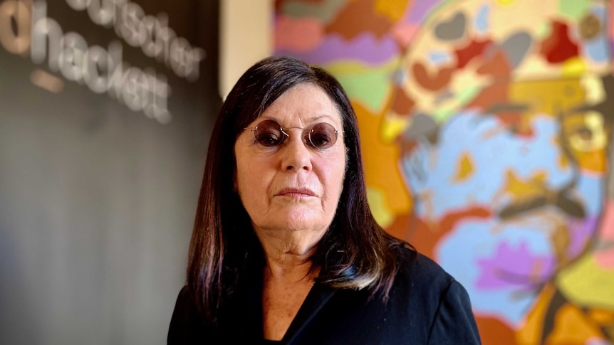 A woman with round, tinted glasses stands in front of a colourful artwork