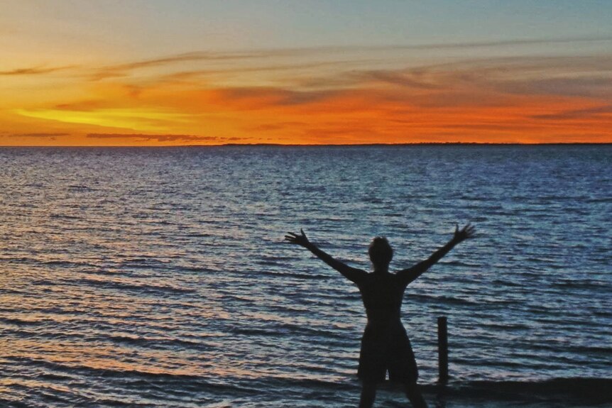 A woman raises her hands in joy with a sun setting over the ocean