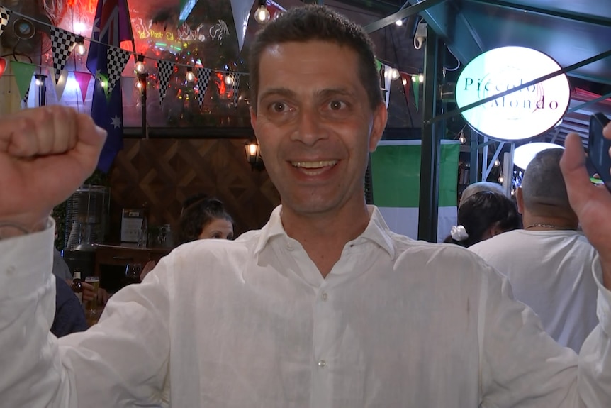 A man wearing a white button up shirt holds both fists in the air and smiles outside a restaurant.