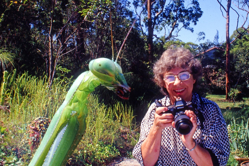 A woman holds a camera up, with a grasshopper in the foreground.