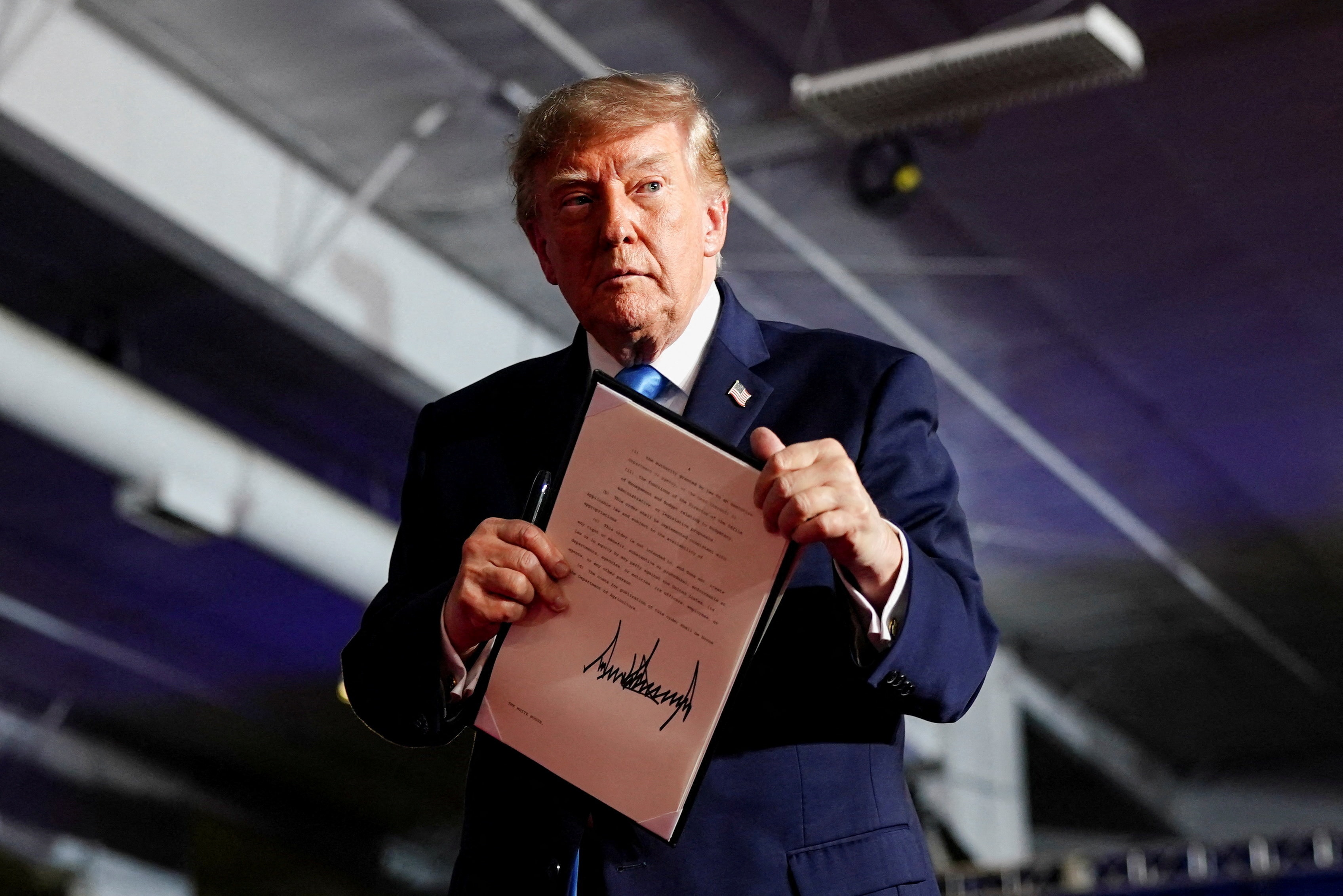 Donald Trump wearing a navy suit and blue tie holding a signed piece of paper in front of him.
