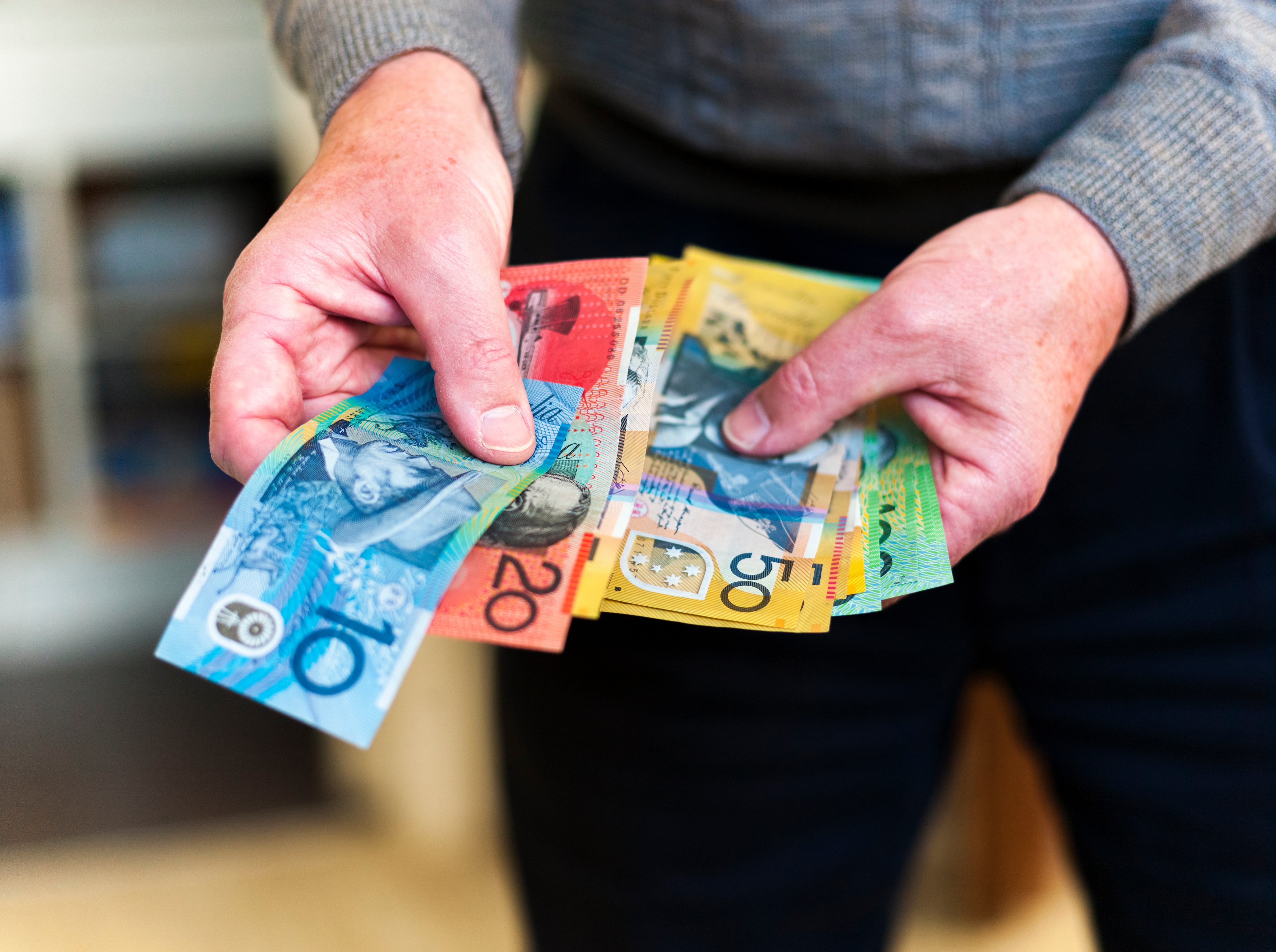 A man wearing a long sleeve jumper holds a wad of Australian currency. There are $10, $20, $50 and $100 notes.