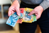 A man wearing a long sleeve jumper holds a wad of Australian currency. There are $10, $20, $50 and $100 notes.