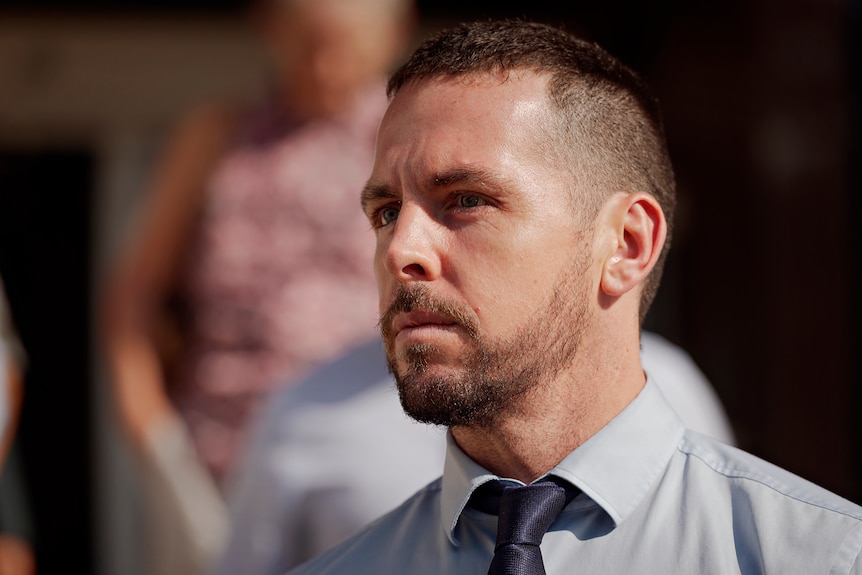 A man in a business shirt and tie looks seriously off the camera.