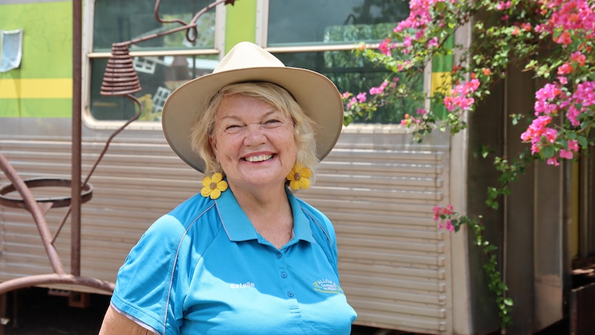 A woman with blonde hair and a wide-brimmed hat smiling with a train carriage behind her