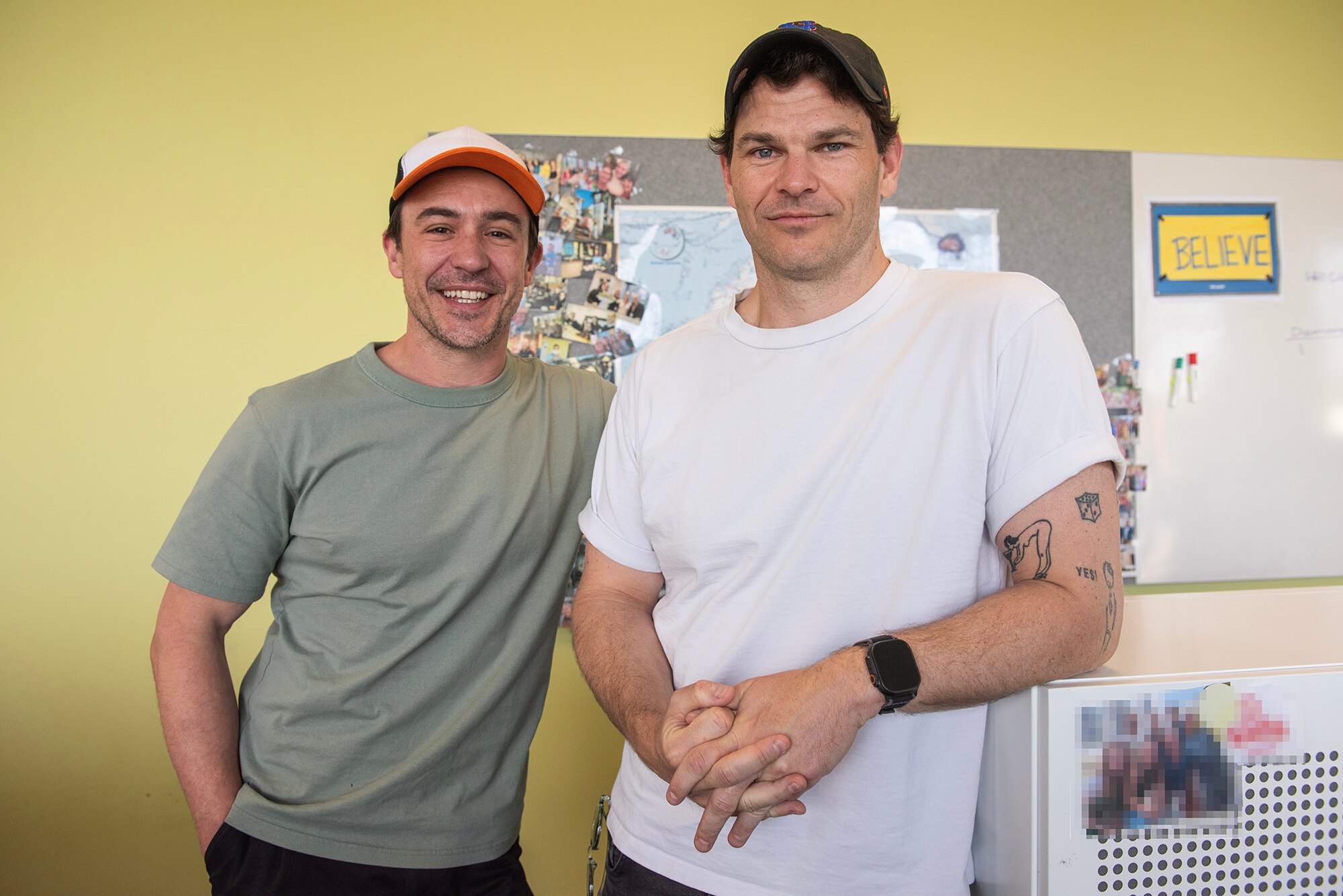 Two young men in hats stand in an office with a yellow wall
