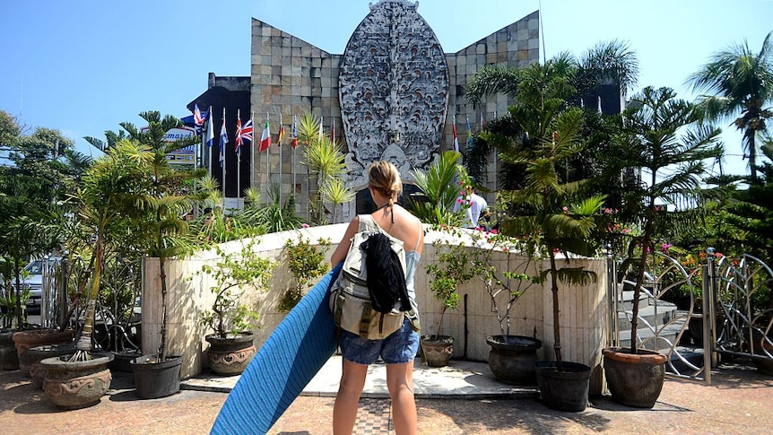 A tourist holding a surfboard stands in front of the Bali bombing memorial in Kuta, Indonesia